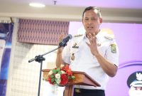 Seorang pejabat Pemerintah Kota Bekasi menyampaikan sambutan dalam sebuah acara resmi, berdiri di belakang podium dengan rangkaian bunga dan menggunakan seragam dinas putih. Ia terlihat menekankan poin penting dengan gestur tangan saat berbicara di depan peserta acara.