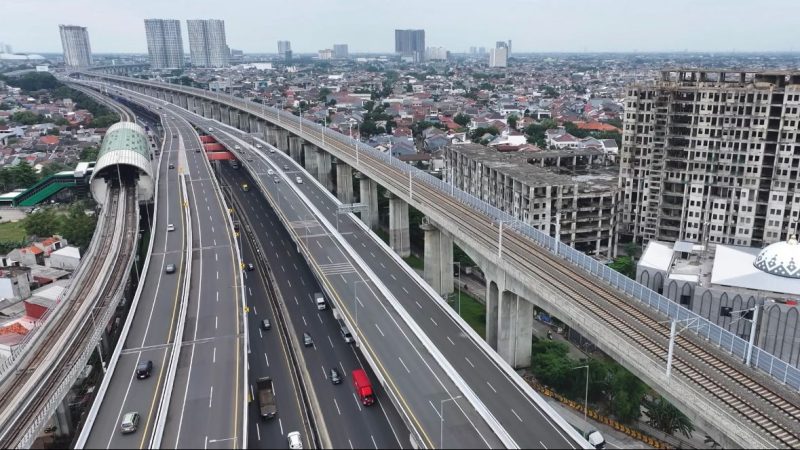 Tampak ruas jalan tol perkotaan yang terintegrasi dengan jalur transportasi massal (LRT) melintasi kawasan permukiman padat dan pusat pembangunan di wilayah Jabodetabek, mencerminkan pengembangan infrastruktur transportasi untuk meningkatkan konektivitas dan mengurai kemacetan perkotaan.