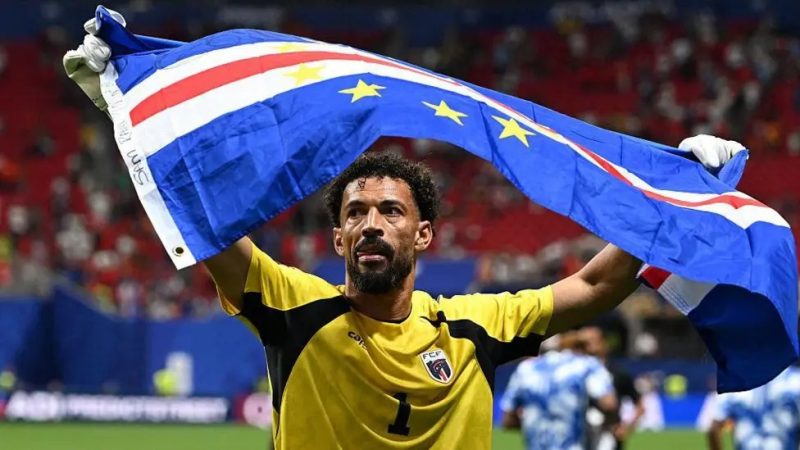 Kiper Tanjung Verde, Vozinha, mengangkat bendera negaranya usai pertandingan melawan Argentina pada babak 32 besar Piala Dunia 2026 di Miami. Meski kalah dramatis 2-3, penampilan heroik Blue Sharks sukses mencuri perhatian dunia sepak bola. (Foto: BBC/Getty Images)