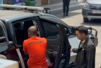Petugas mengawal seorang warga negara Nigeria berinisial OCO yang mengenakan pakaian bertuliskan “DETENI” saat hendak dimasukkan ke dalam kendaraan. OCO ditetapkan sebagai tersangka dugaan tindak pidana keimigrasian setelah tercatat overstay selama 3.073 hari atau lebih dari delapan tahun.