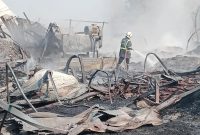 Petugas pemadam kebakaran melakukan pendinginan di lokasi kebakaran. Sejumlah bangunan dan material tampak hangus terbakar, sementara asap masih mengepul di area kejadian.